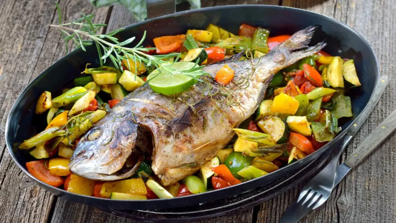 Dorada z piekarnika podana z kolorowymi warzywami: papryką, cukinią i pomidorami, udekorowana rozmarynem i limonką.