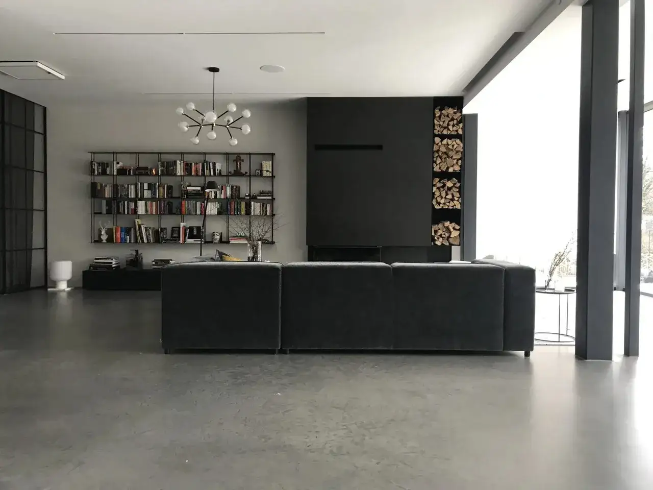 Nowoczesny salon z szarą, gładką posadzką betonową w domu. Wygodna, ciemnoszara kanapa, regał z książkami i czarny kominek.