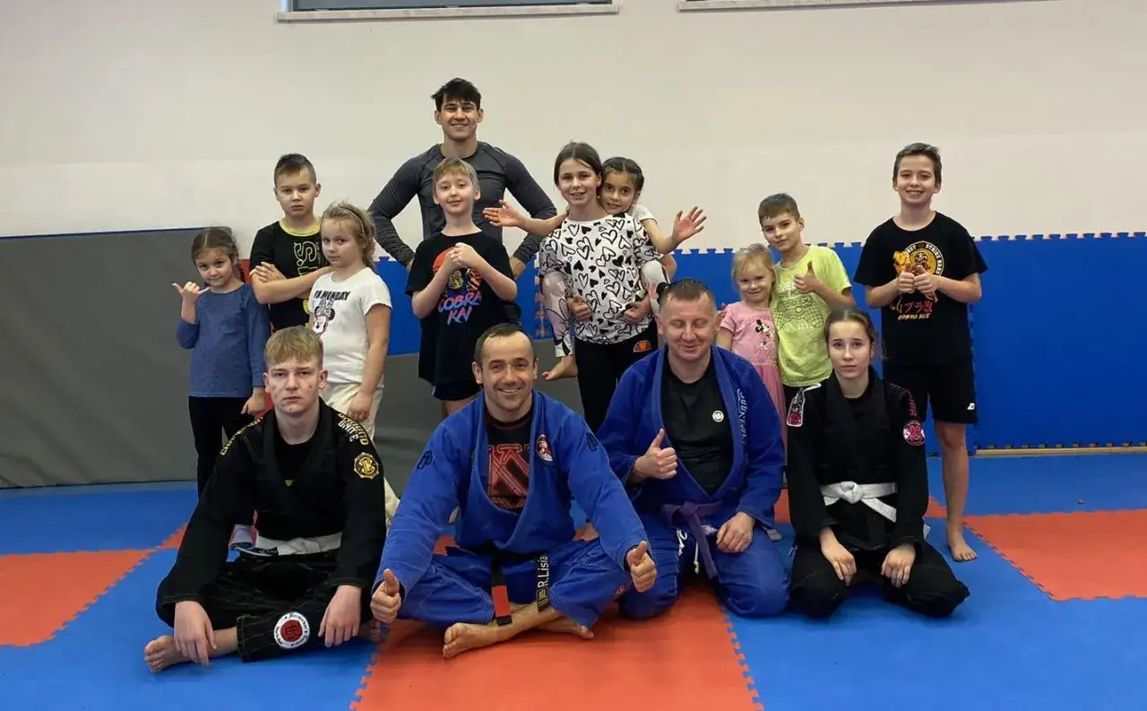 Grupa dzieci i trener&oacute;w na macie po treningu mma dla dzieci. Wszyscy uśmiechnięci, niekt&oacute;rzy pokazują kciuki w g&oacute;rę.