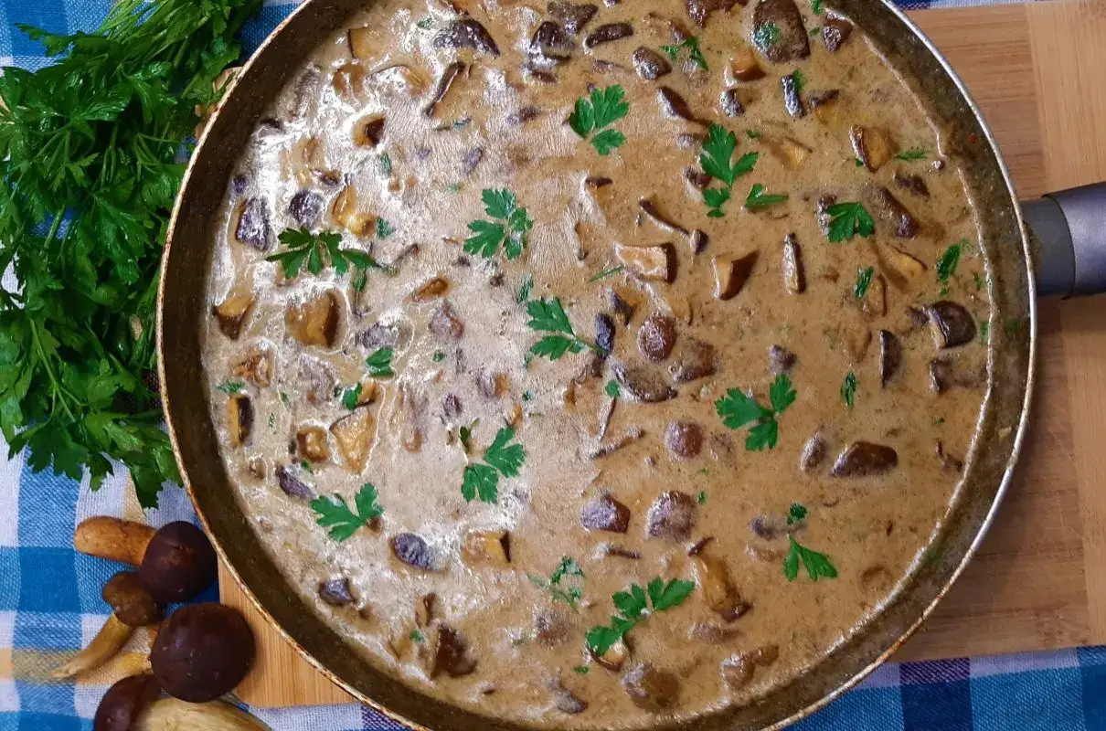 Pyszny, kremowy sos grzybowy w patelni, udekorowany natką pietruszki. Obok świeże grzyby i pęczek zieleniny.