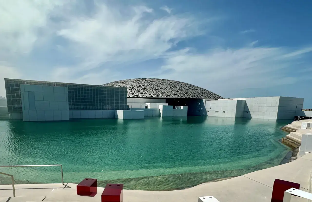 Luwr Abu Zabi z charakterystyczną kopułą, otoczony turkusową wodą. Nowoczesna architektura w słońcu.