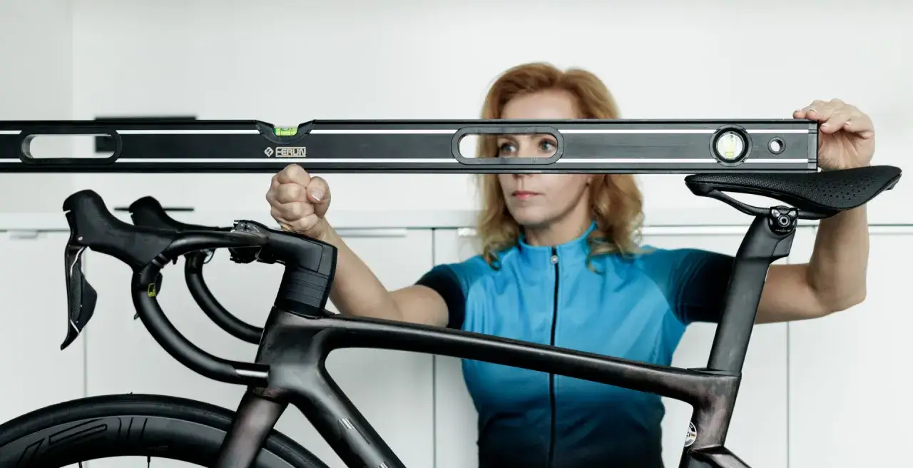 Kobieta w niebieskiej koszulce kolarskiej używa poziomicy do precyzyjnego bike fittingu, sprawdzając ustawienie kierownicy i siodełka.