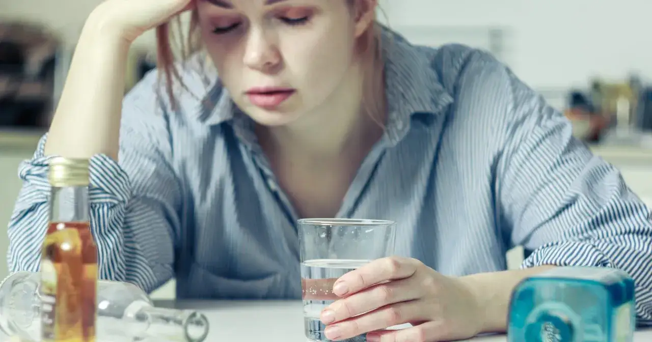 Zmęczona kobieta z bólem głowy pije wodę, szukając sposobu, jak szybciej wytrzeźwieć. Obok leżą puste butelki.