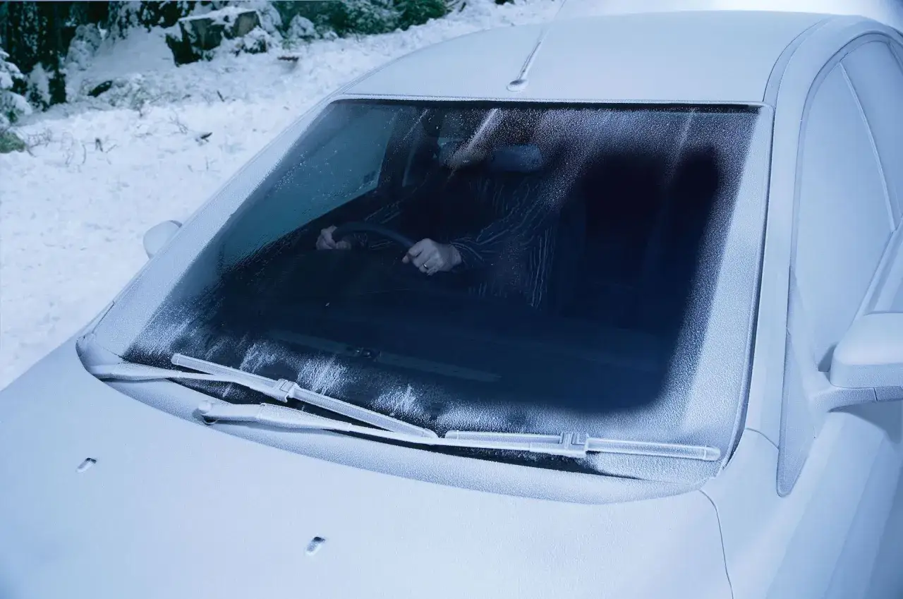 Zamarznięta przednia szyba samochodu. Kierowca w środku, pr&oacute;buje uruchomić podgrzewanie przedniej szyby, by ruszyć w mroźny dzień.