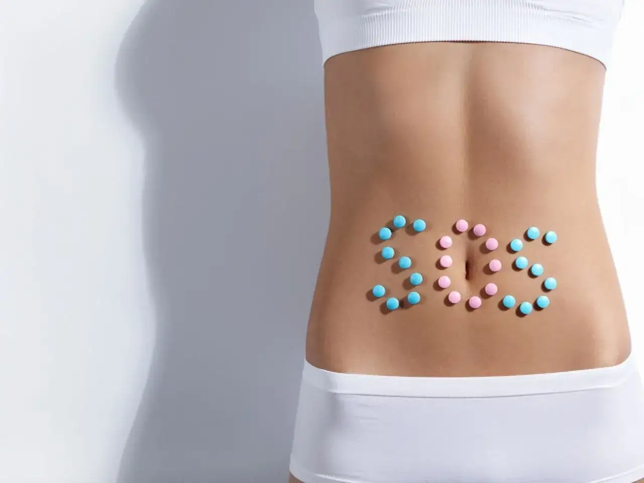 Niebieskie i różowe tabletki ułożone na brzuchu w napis SOS. Szukasz domowych sposobów na ból brzucha?