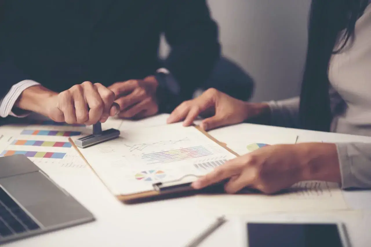 Dwie osoby analizują wykresy i dokumenty. Jedna z nich przybija pieczątkę, co pokazuje, ile są ważne dokumenty do sprzedaży działki.