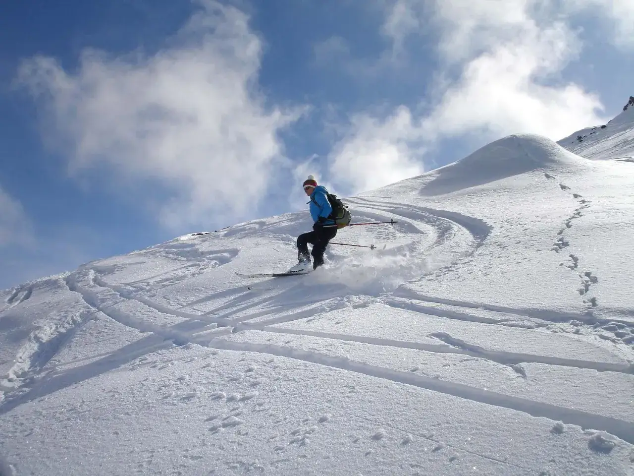 Narciarz zjeżdża po ośnieżonym stoku, zostawiając ślady. W tle błękitne niebo z chmurami. Wyciąg narciarski Karpacz w oddali.