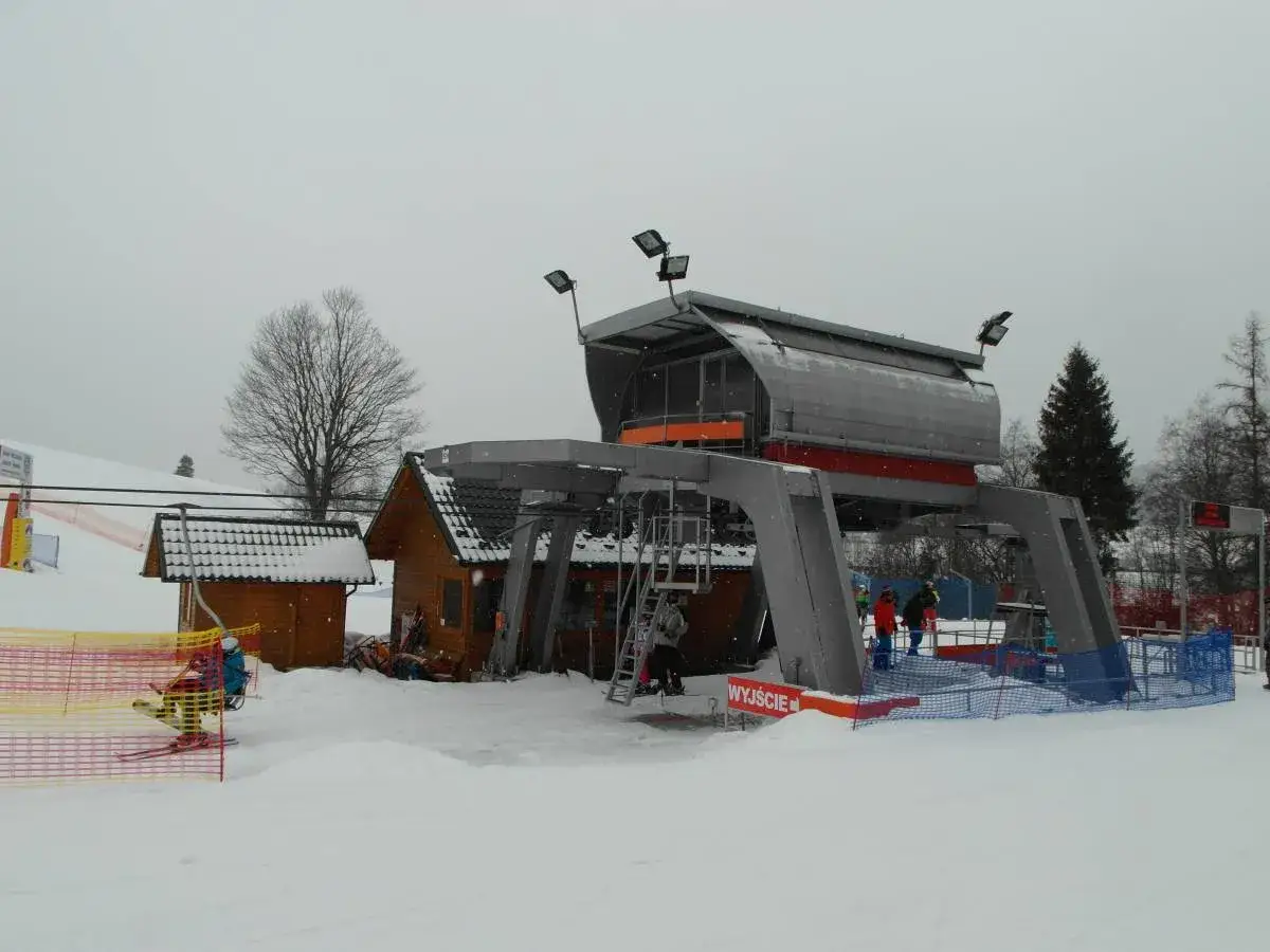 Stacja narciarska z drewnianymi budynkami i nowoczesnym wyciągiem. Na stoku widać narciarzy i ogrodzenie.