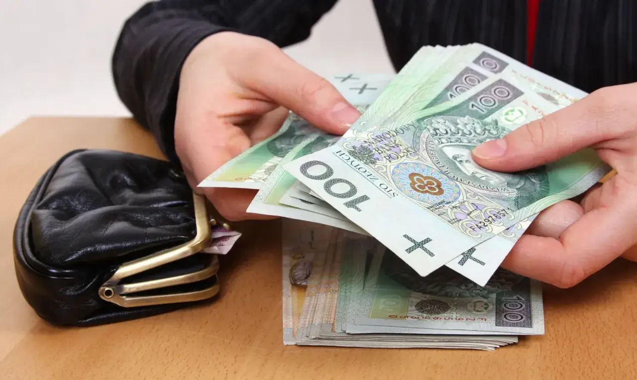 Dłonie liczą banknoty 100 zł. Czy to już kwota, ile wynosi 13 emerytura? Obok leży czarny portfel.