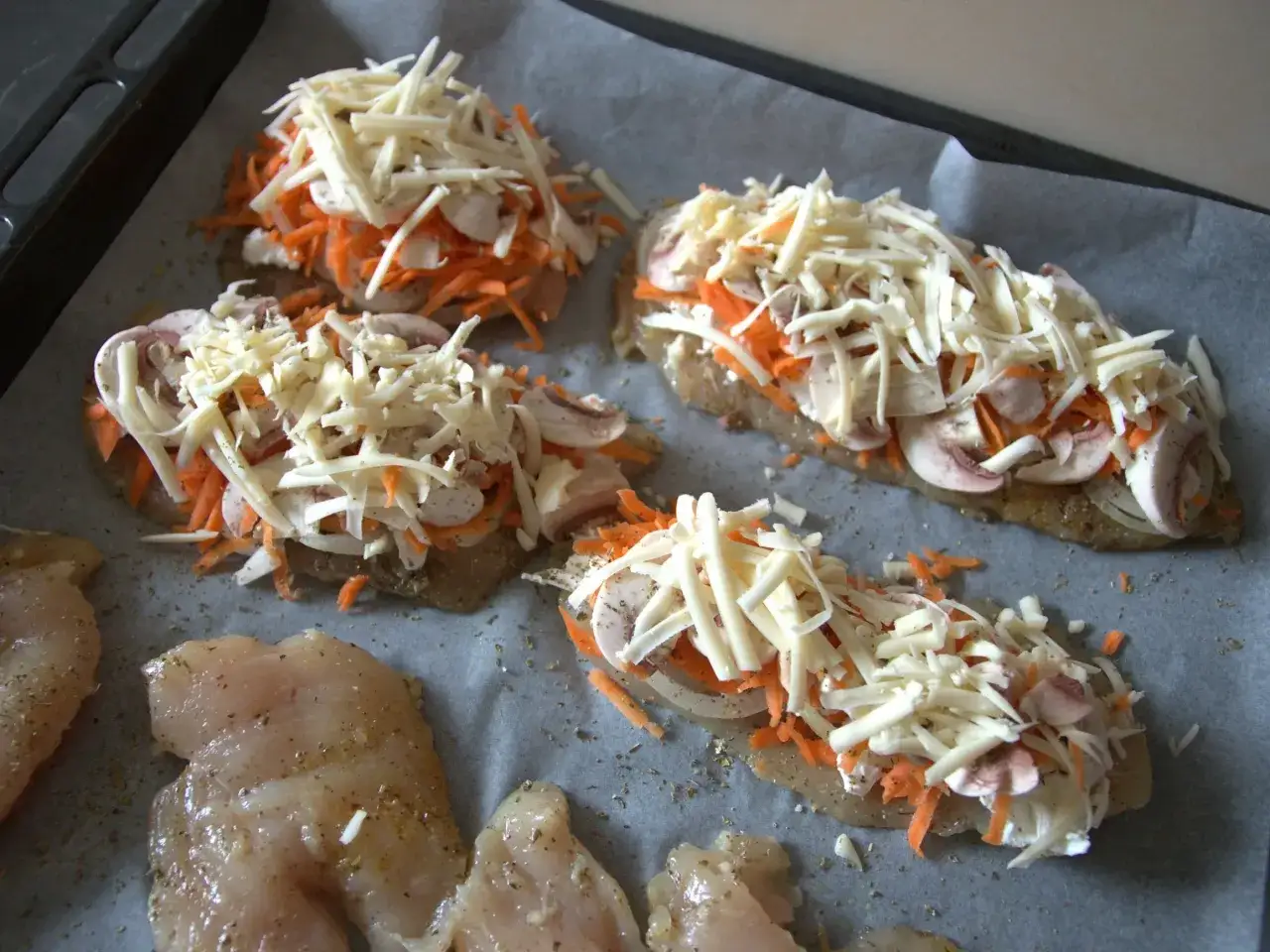 Na blasze wyłożonej papierem do pieczenia leżą surowe kawałki soczystej piersi z kurczaka, przygotowane do zapieczenia z warzywami i serem.