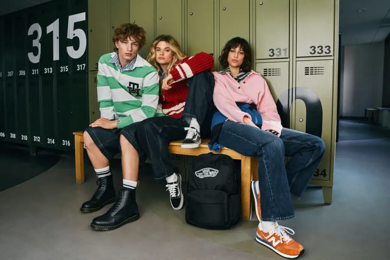 Tr&oacute;jka młodych ludzi w luźnych, modnych ubraniach, prezentująca styl ubioru młodzieży. Siedzą na ławce przed zielonymi szafkami.