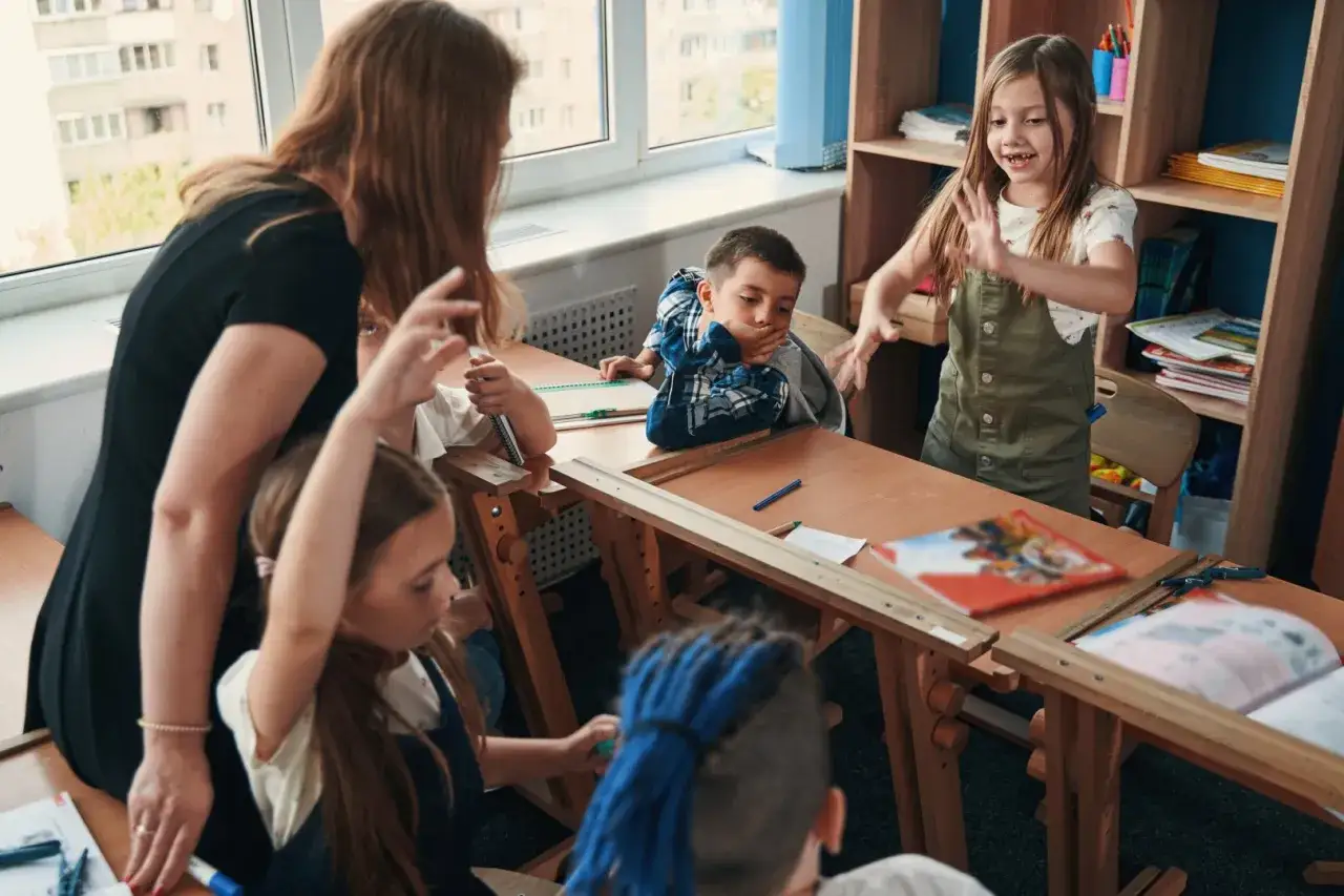 Nauczycielka omawia rozporządzenie w sprawie kwalifikacji nauczycieli z uczniami. Dzieci aktywnie uczestniczą w lekcji, podnosząc ręce i zadając pytania.