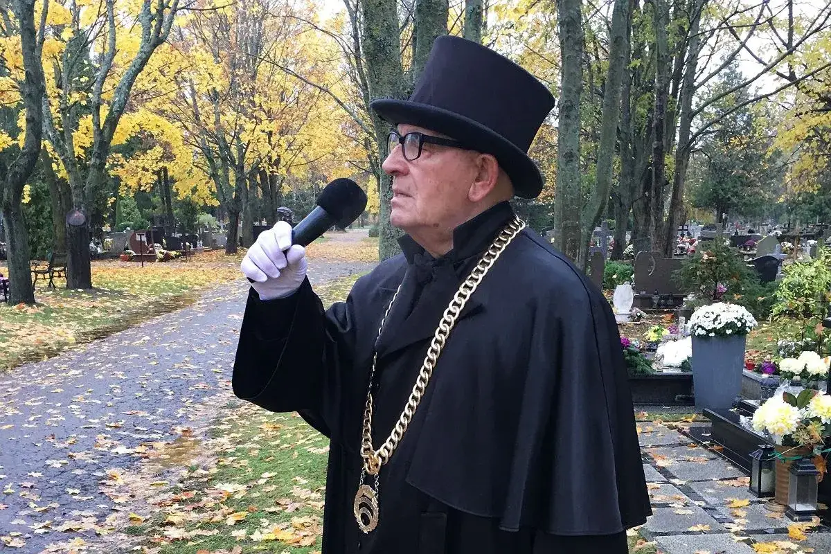 Mężczyzna w cylindrze i todze z łańcuchem przemawia przez mikrofon na cmentarzu. Długość ceremonii, ile trwa pogrzeb, zależy od wielu czynników.