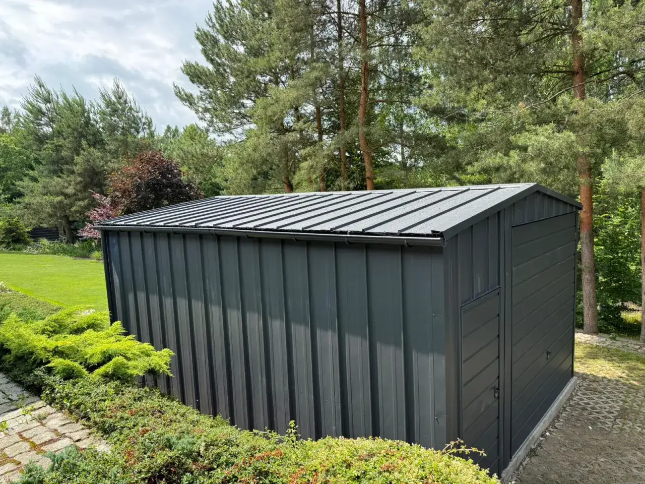 Nowy domek narzędziowy z ciemnoszarą blachą na rabek, otoczony zielenią i drzewami.
