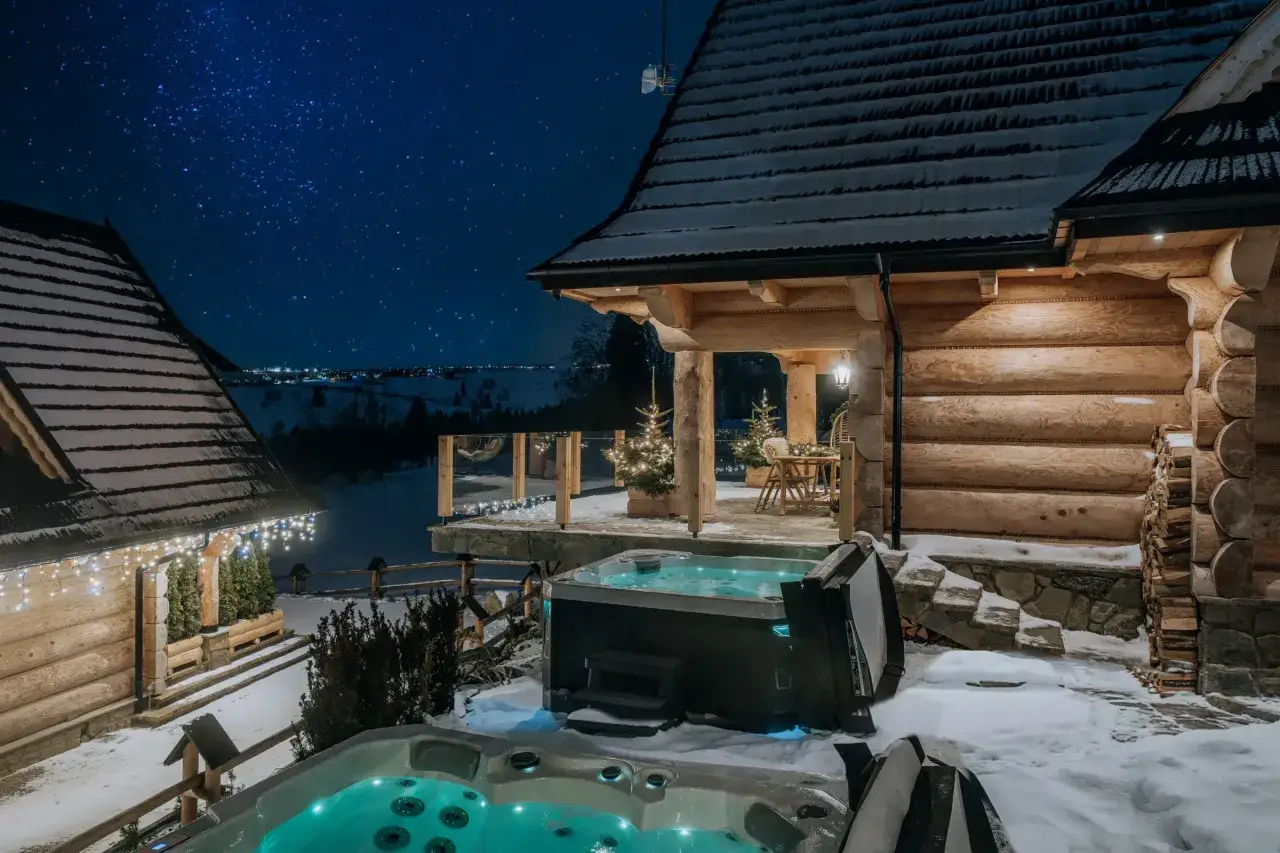 Nocne niebo nad Bukowiną Tatrzańską. Drewniany domek z oświetleniem i gorącymi baliami w śniegu.