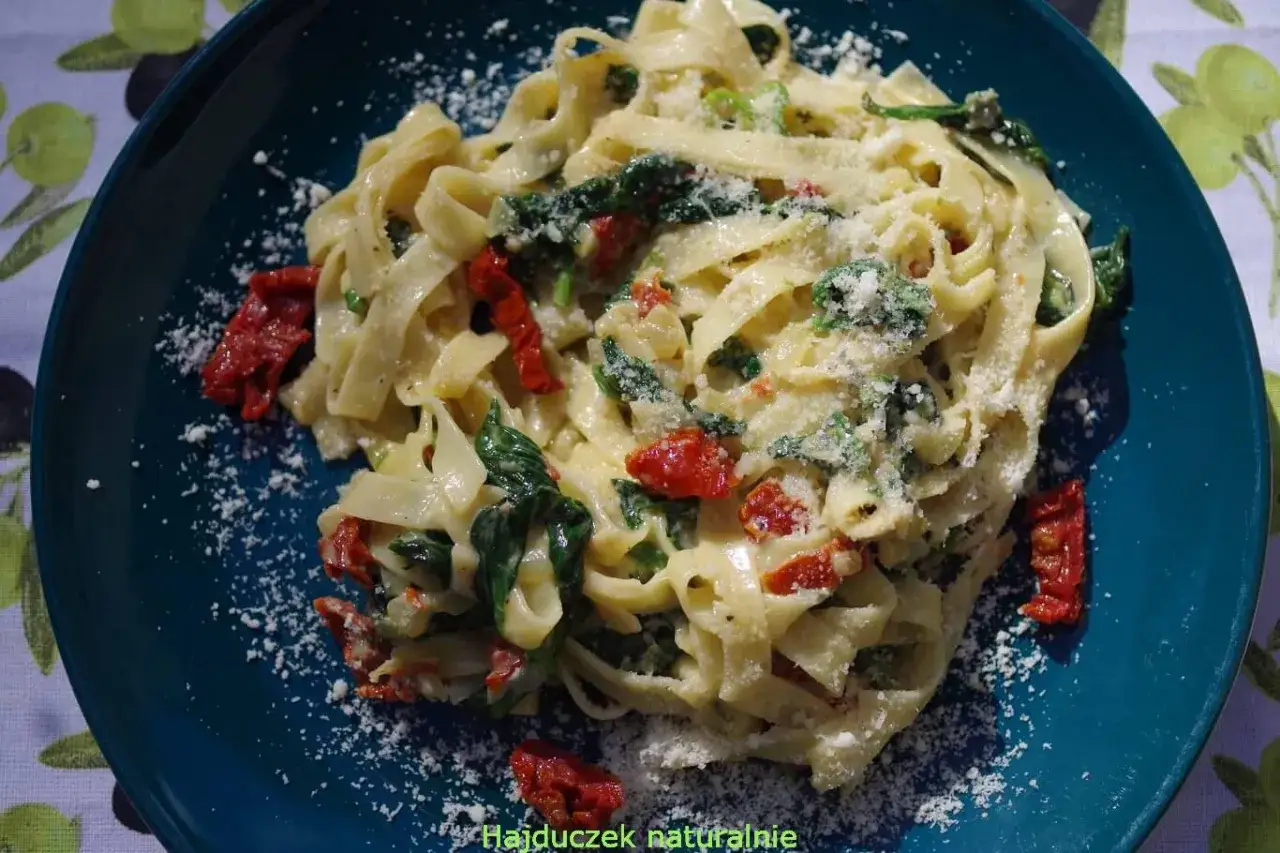 Pyszny makaron ze szpinakiem i suszonymi pomidorami, posypany parmezanem, na talerzu z motywem oliwnym.
