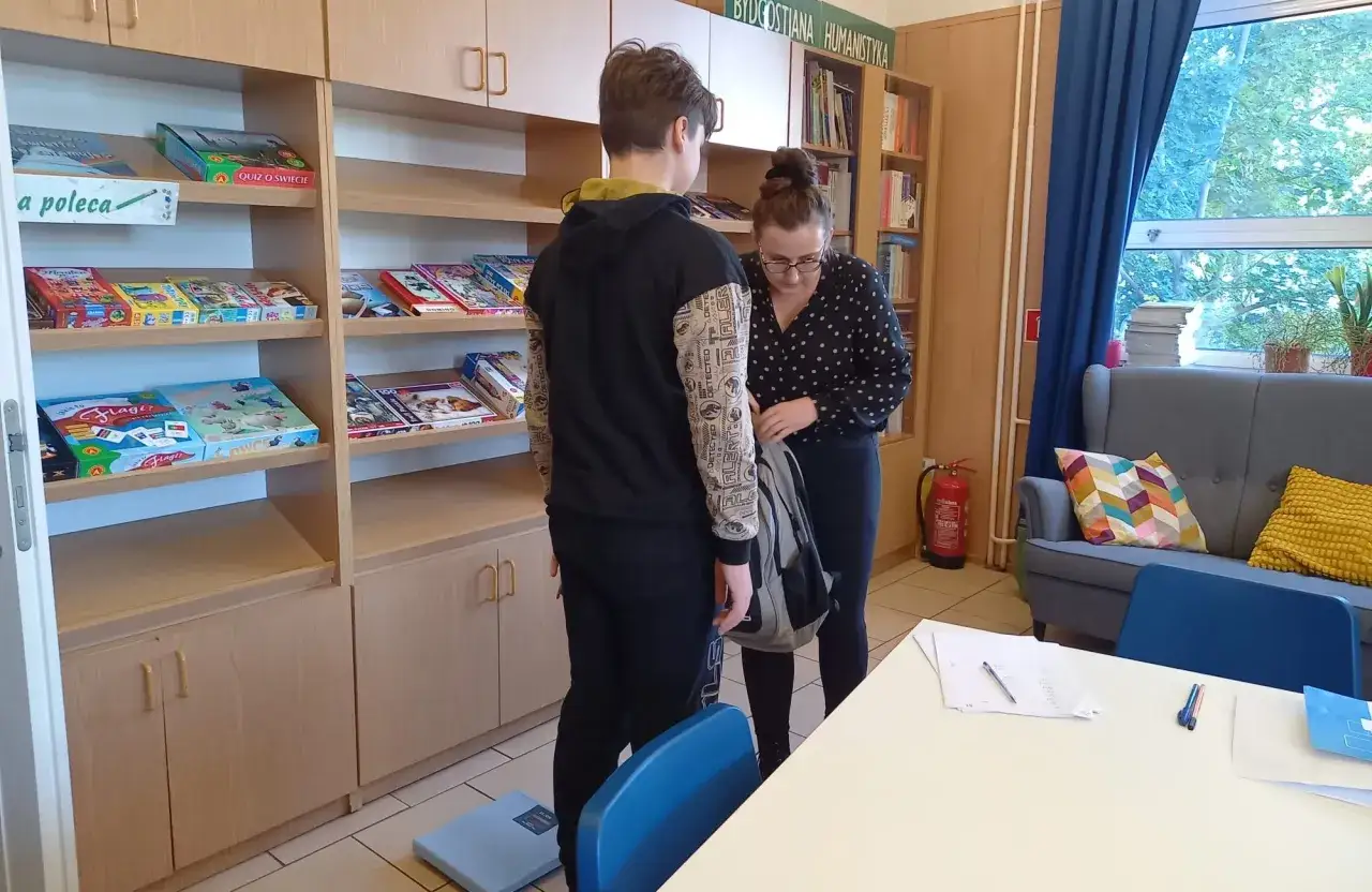 W szkole, przy regałach z grami i książkami, młodzi ludzie przeglądają zawartość szafek.