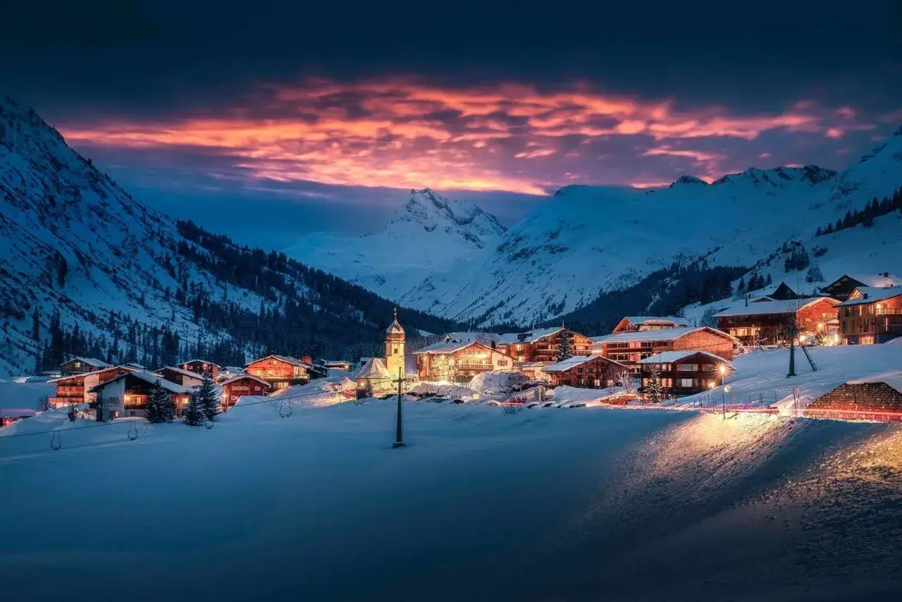 Piękne zimowe zdjęcia g&oacute;rskiej wioski o zmierzchu. Oświetlone domy i kości&oacute;ł toną w śniegu, a niebo rozświetla purpurowy zach&oacute;d słońca.
