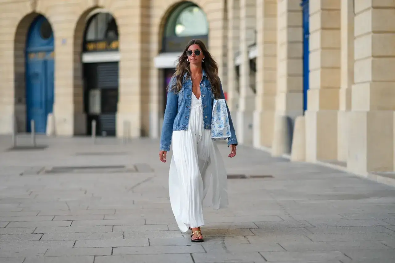 Kobieta w zwiewnej białej sukience i jeansowej kurtce, z torbą w ręku, spaceruje po brukowanym chodniku.