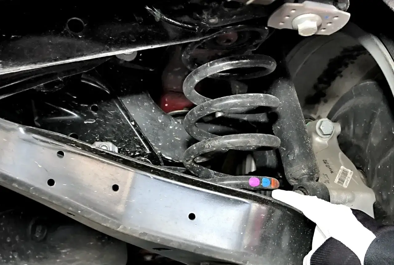 Widok z dołu na zawieszenie samochodu, gdzie trwa wymiana sprężyn prz&oacute;d. Widać amortyzator i sprężynę z kolorowymi oznaczeniami.