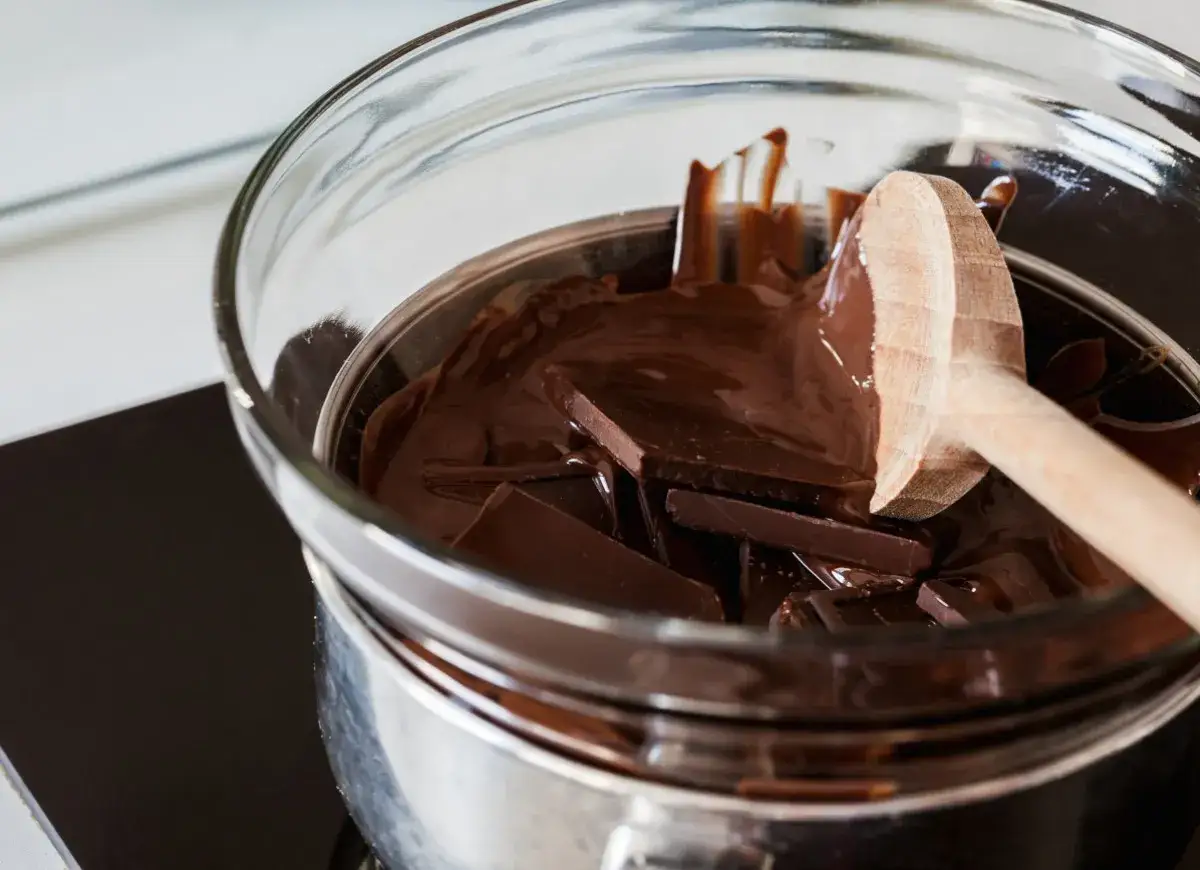 Czekolada w kawałkach topi się w szklanej misce nad garnkiem z wodą. Drewniana łyżka miesza płynną masę, pokazując, jak rozpuścić czekoladę w kąpieli wodnej.