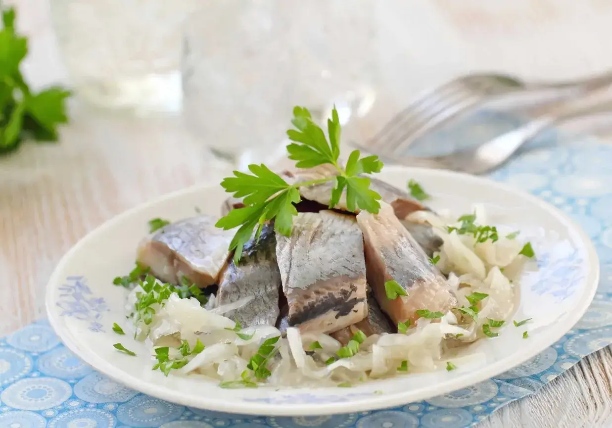 Jak zrobić dobre śledzie po wiejsku? Na talerzu ułożone kawałki śledzia z cebulką i natką pietruszki.