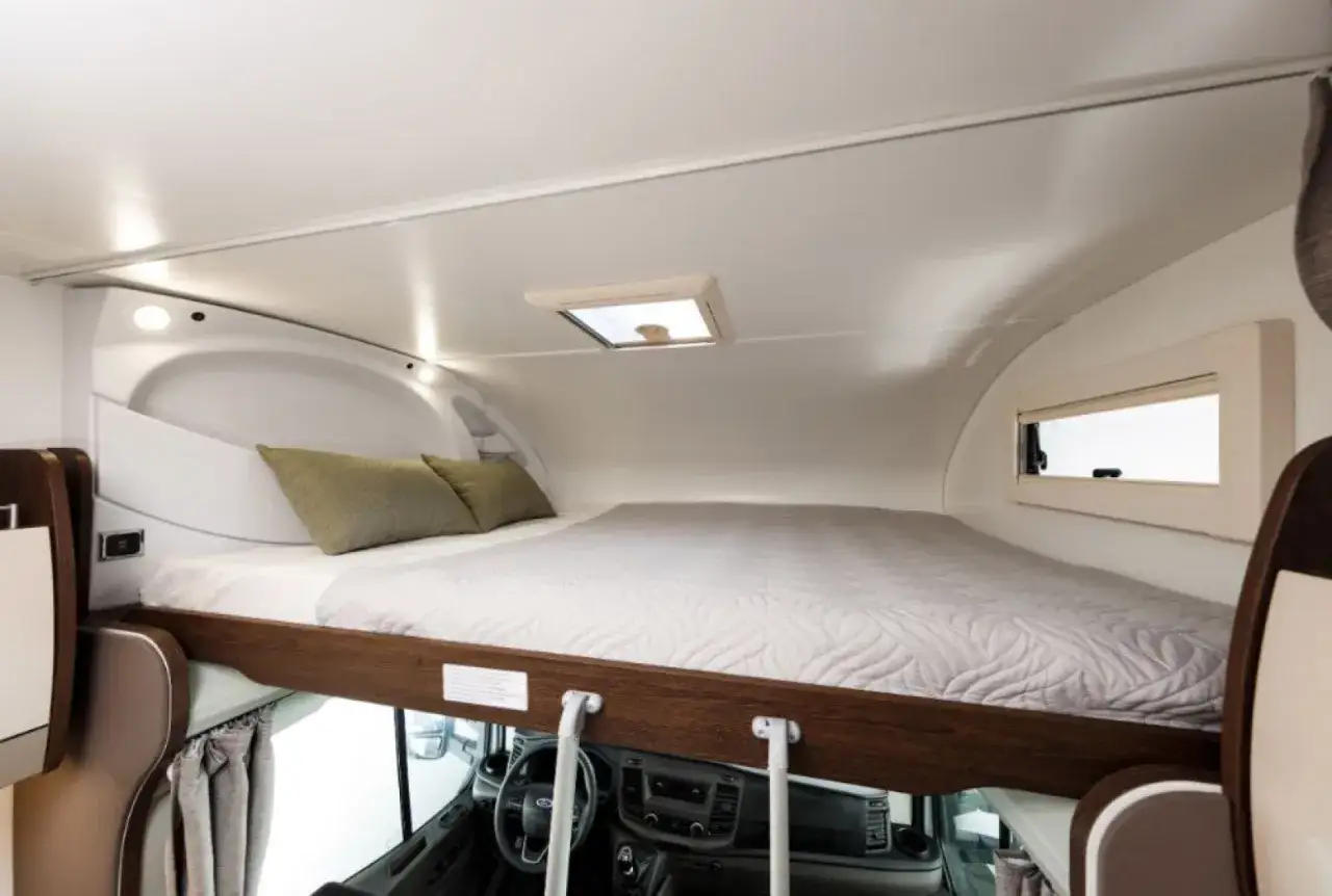Wygodne łóżko nad kabiną kierowcy w kamperze alkowa. Dwie poduszki i okno dachowe zapewniają komfort.