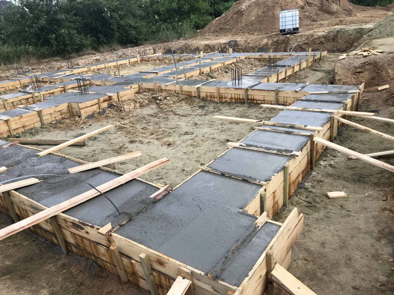 Budowa fundamentów: beton wylewany do drewnianych szalunków, zbrojenie wystaje z betonu.