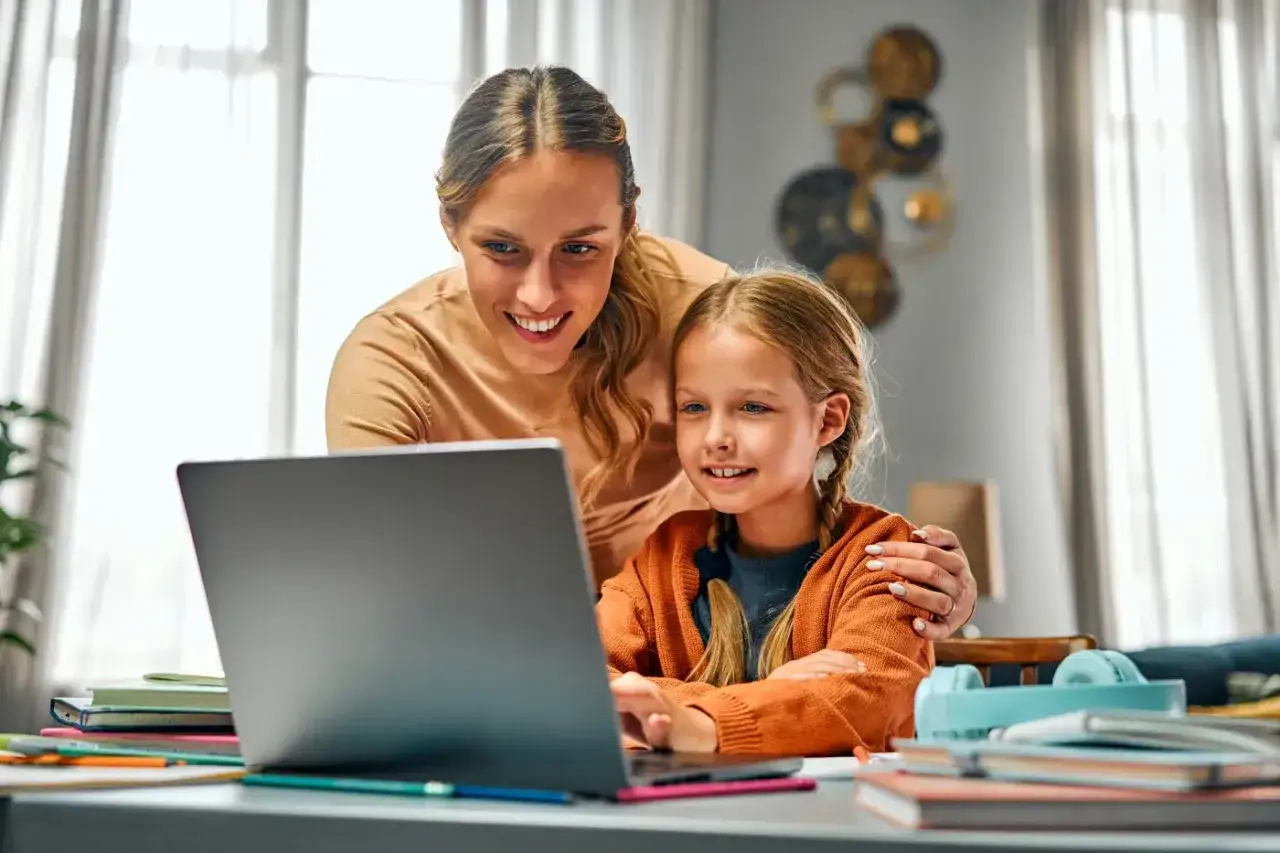 Uśmiechnięta mama wspiera córkę w nauce przy laptopie. To przykład troskliwej opieki na dziecko podczas odrabiania lekcji.