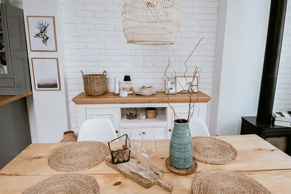 Drewniany st&oacute;ł z okrągłymi podkładkami, wazonem i szkłem. Komoda z koszem i dekoracjami. Styl rustykalny nowoczesny.
