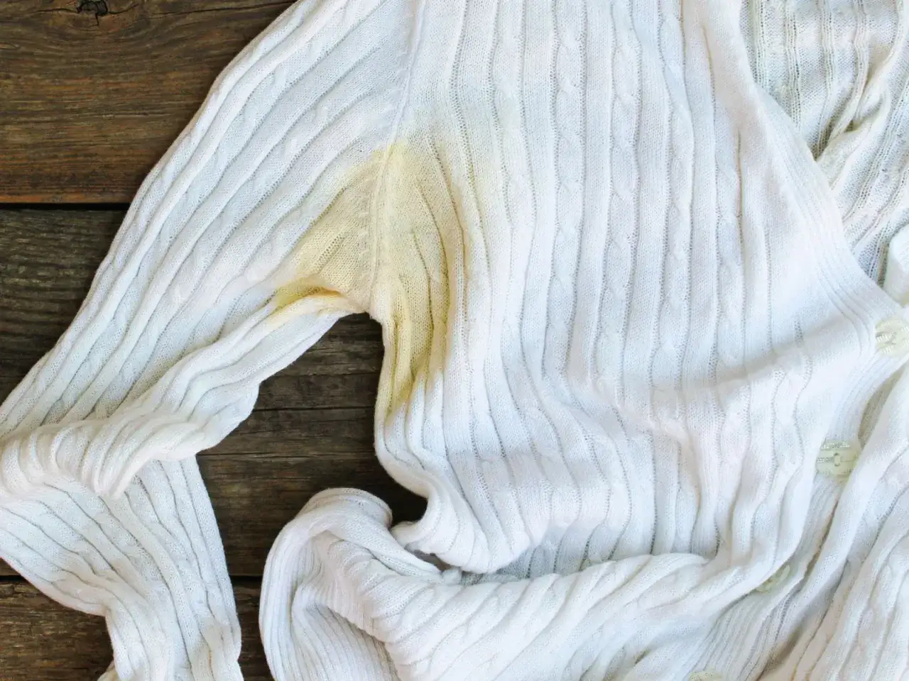 Biały sweter z warkoczowym splotem ma widoczne ż&oacute;łte plamy pod pachą, świadczące o trudnych do usunięcia zabrudzeniach na białych ubraniach.