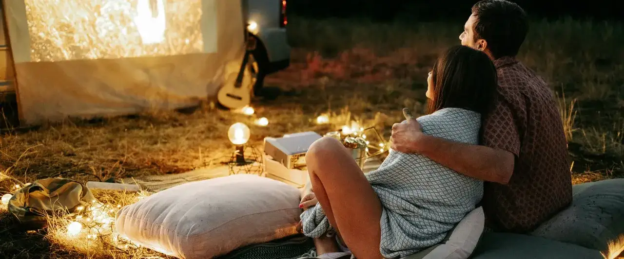 Para 100 pomysł&oacute;w na randkę: para przytulona ogląda film pod gołym niebem, otoczona lampkami i poduszkami.