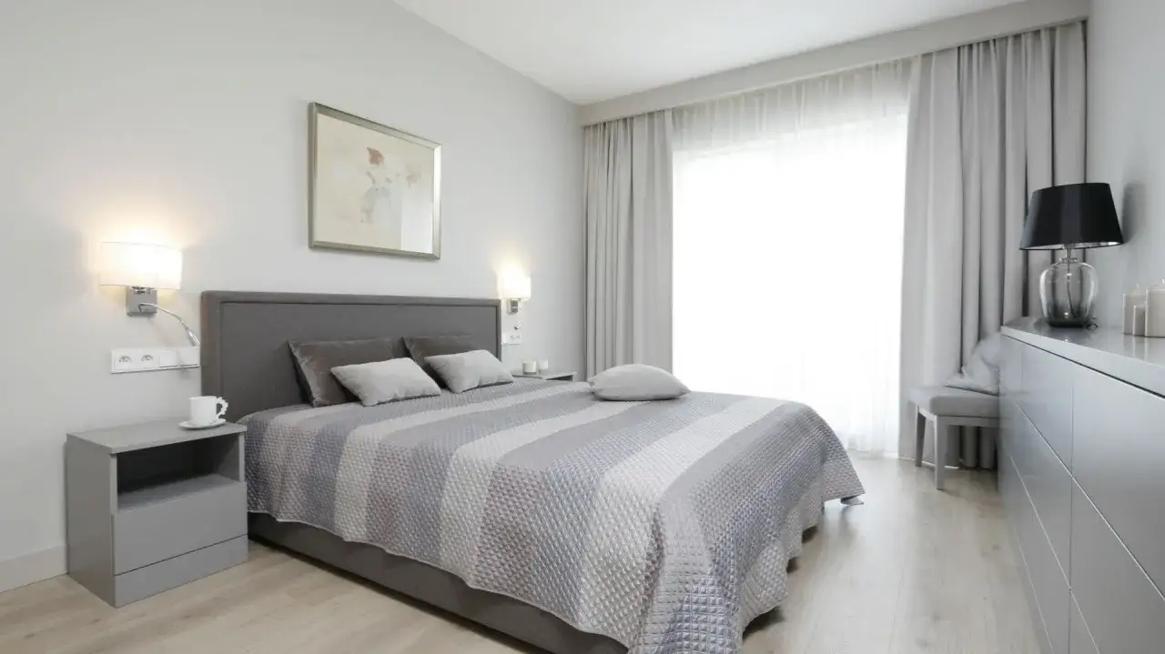 Przytulna sypialnia z kinkietami nad ł&oacute;żkiem. Szare tekstylia i jasne drewno tworzą spokojną atmosferę.