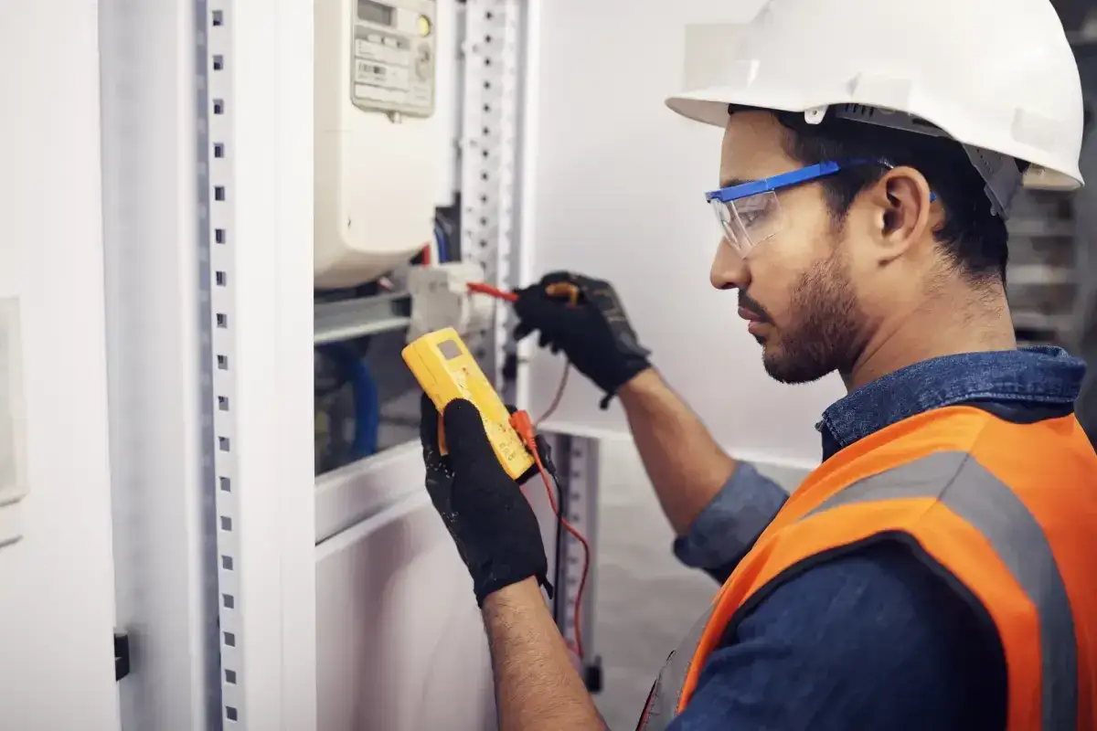 Elektryk w kasku i okularach ochronnych sprawdza instalację elektryczną za pomocą miernika. To część obowiązkowych przeglądów budynków.