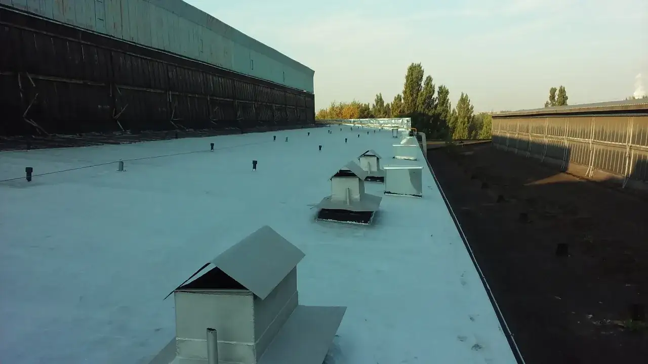Srebrne kominki wentylacyjne na białym dachu przemysłowym, otoczone przez drzewa i budynki.