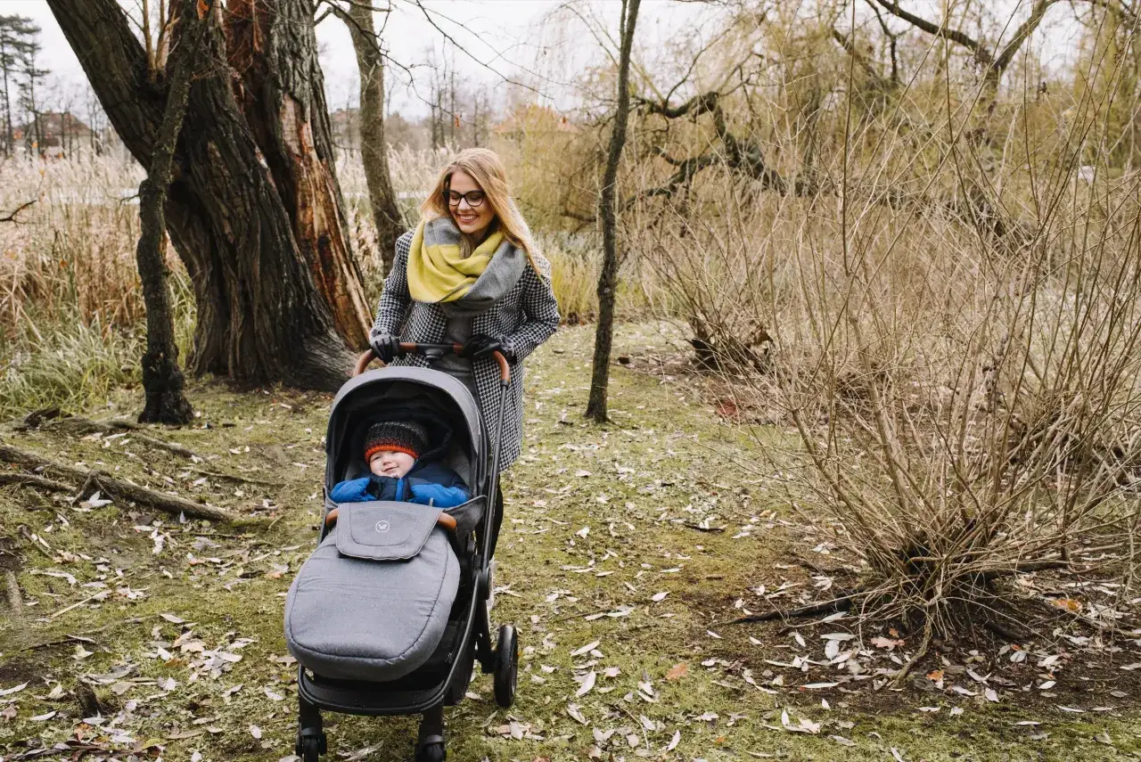 Mama z uśmiechem pcha w&oacute;zek z 2-letnim dzieckiem. Idealny spacer, nawet jeśli nie gdzie w g&oacute;ry z dzieckiem 2 letnim.