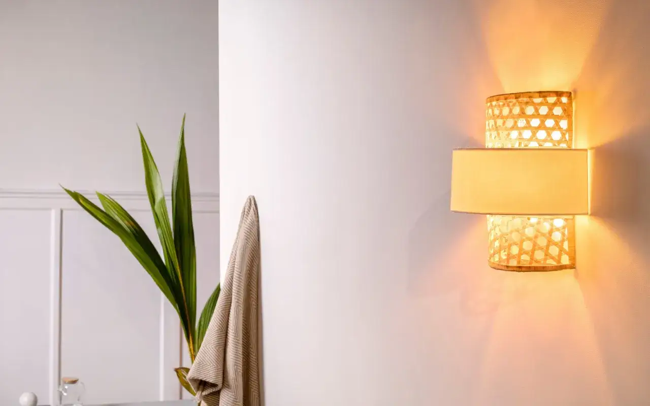 Kinkiet z plecionki rattanowej tworzy ciepłe podświetlenie ściany od dołu, rzucając subtelne cienie. Obok zielone liście rośliny.