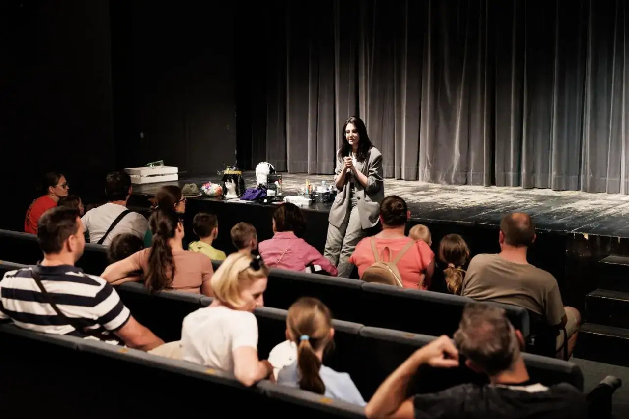 Widownia w teatrze słucha opowieści o tym, jak twoja twarz brzmi znajomo. Na scenie stoją peruki i rekwizyty.