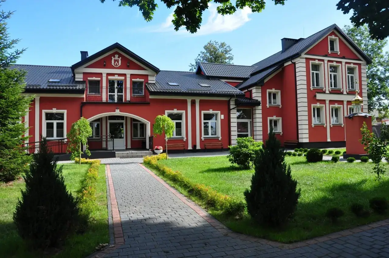 Jasnoczerwony budynek z białymi zdobieniami, otoczony zielenią. To mój dom opieki, gdzie czuję się bezpiecznie i komfortowo.