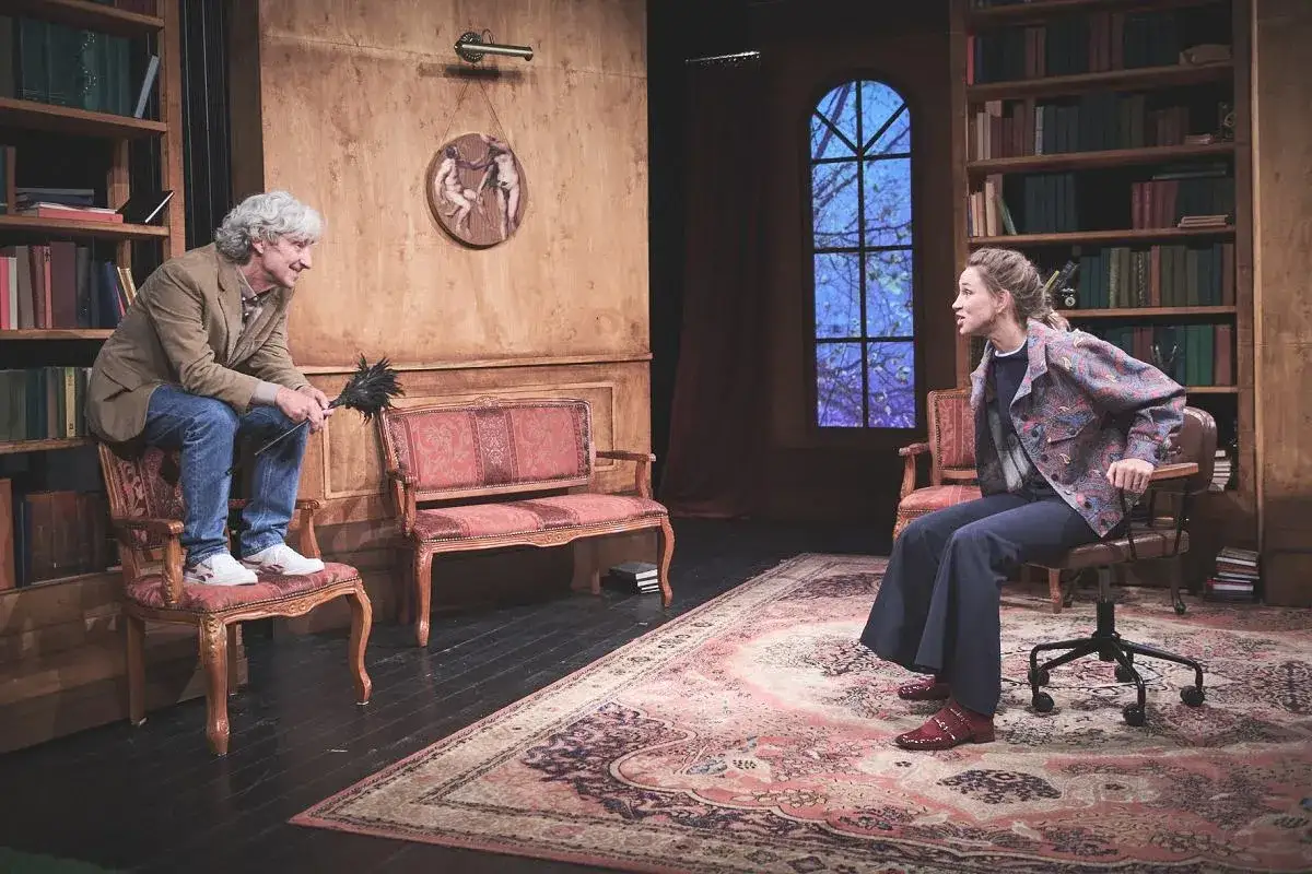 Scena teatralna: mężczyzna z miotełką siedzi na krześle, kobieta na obrotowym fotelu. Edukacja Rity w bibliotece.