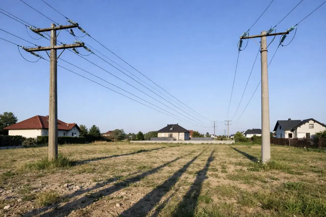 Dwa słupy energetyczne z przewodami wysokiego napięcia 110 kV, nad pustą działką z cieniami domów w tle. Zachowanie bezpieczna odległość od linii wysokiego napięcia 110 kv jest kluczowe.