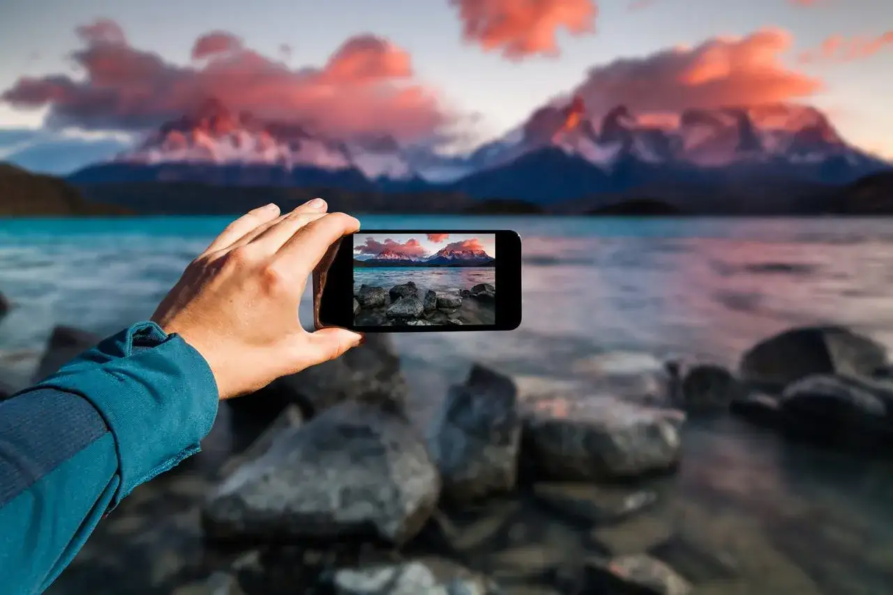 Dłoń trzyma smartfon, na kt&oacute;rym widać zdjęcie g&oacute;r i jeziora. To przykład, jak fotografia obliczeniowa pozwala uchwycić piękno natury.