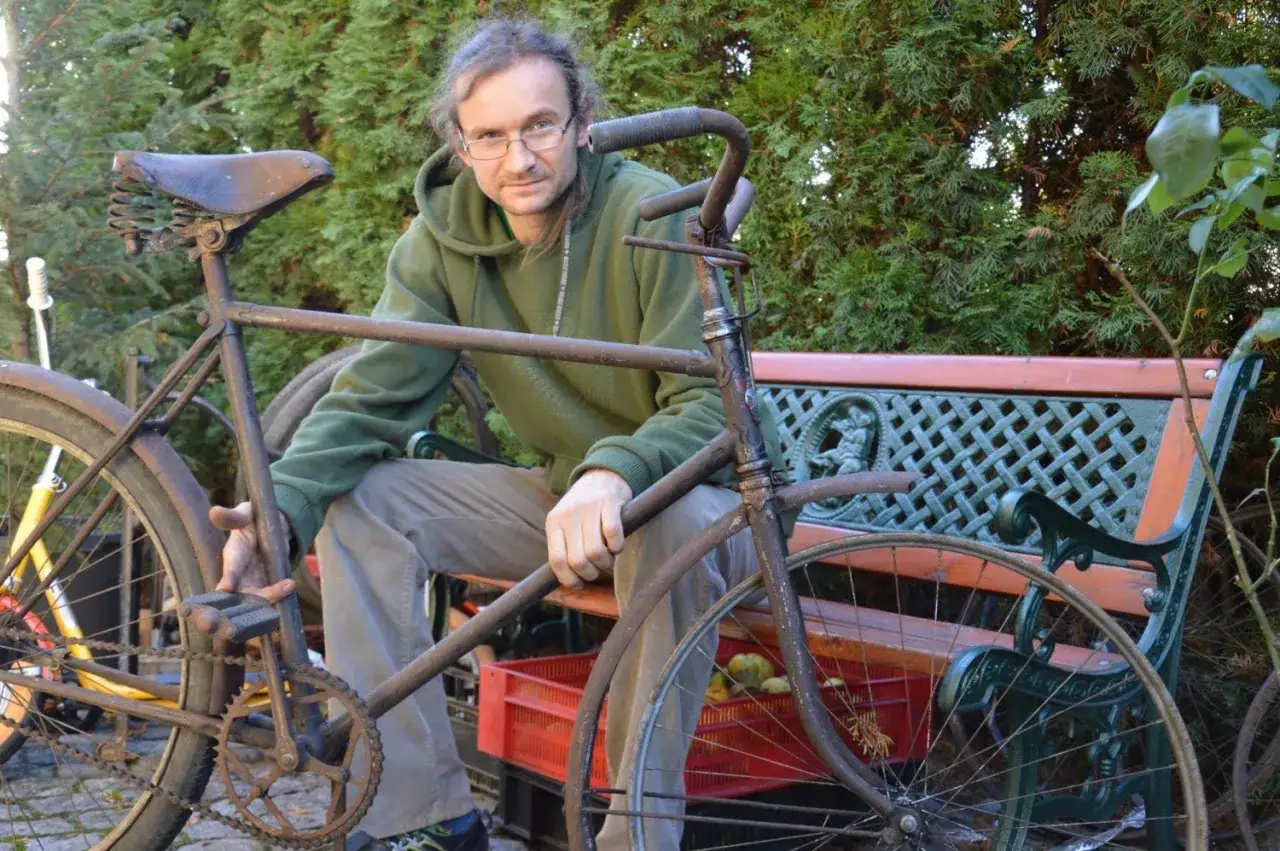 Mężczyzna w zielonej bluzie siedzi obok starego, zardzewiałego roweru. Zastanawia się, gdzie wyrzucić stary rower, który wygląda jak relikt przeszłości.