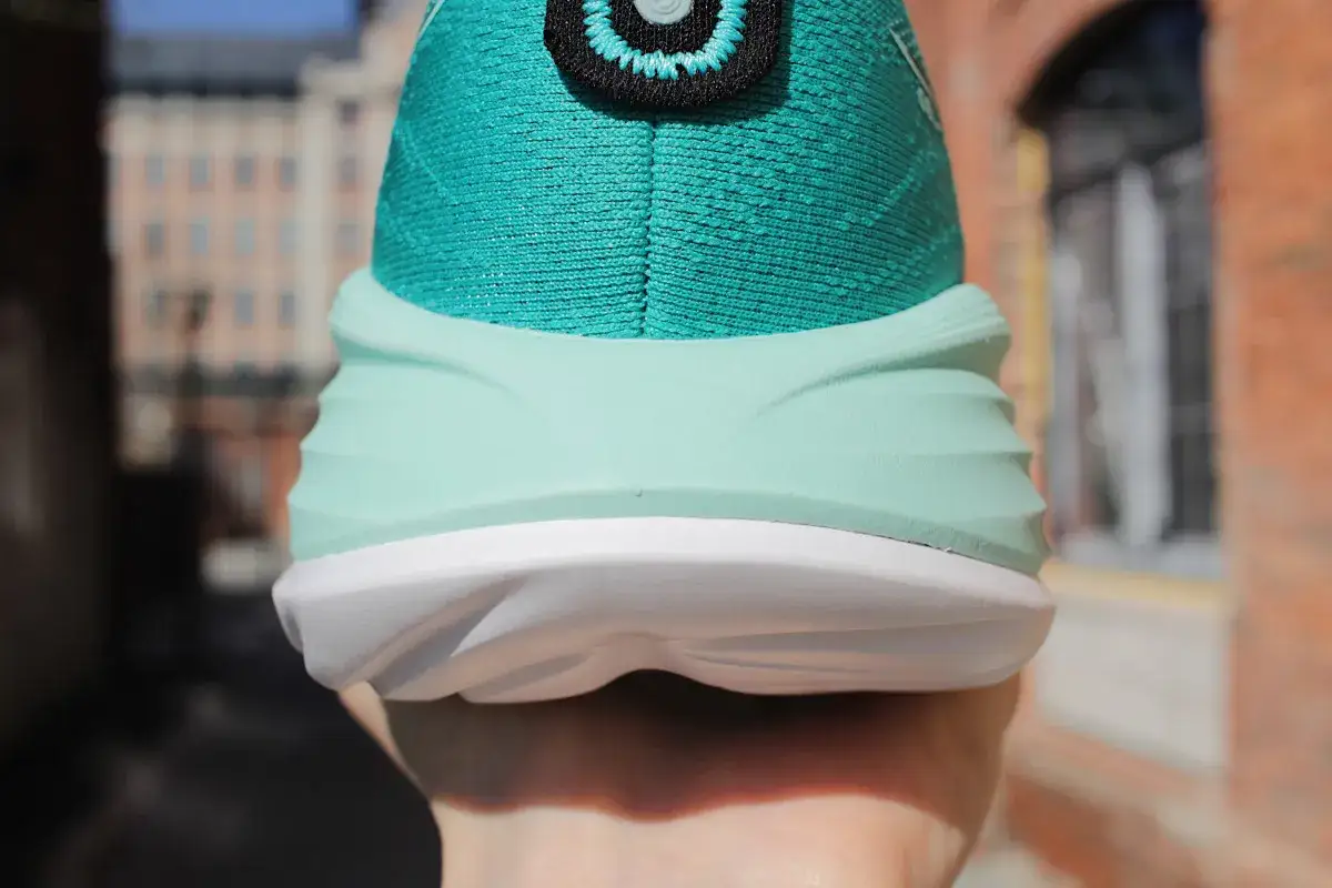 Turkusowe buty supinujące z białą podeszwą, trzymane w dłoni na tle ceglanej ściany i budynk&oacute;w.