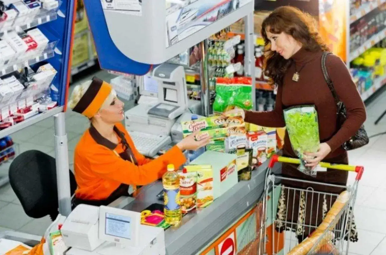 Kasa w sklepie spożywczym, gdzie kasjerka obsługuje klientkę. Obrazek może sugerować, ile zarabia właściciel małego sklepu spożywczego po odliczeniu podatków.