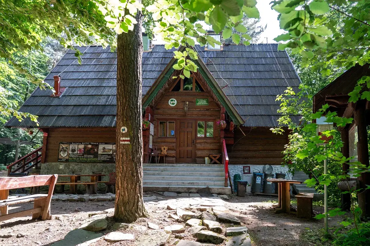 Drewniana bac&oacute;wka PTTK w Bartnem otoczona zielenią. Przed schodami ławki i stoły, a obok drzewo z oznaczeniem szlaku.