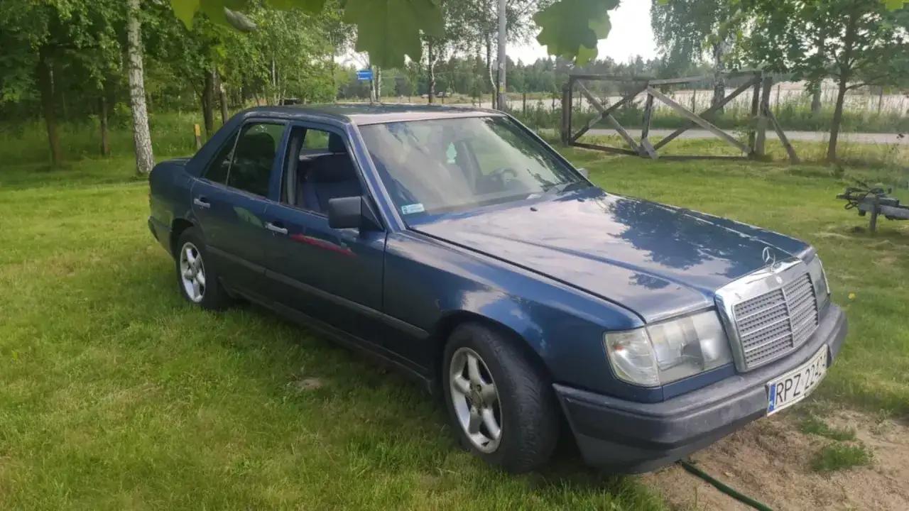 Niebieski Mercedes W124 na trawie, z widokiem na jezioro i drewniane ogrodzenie.