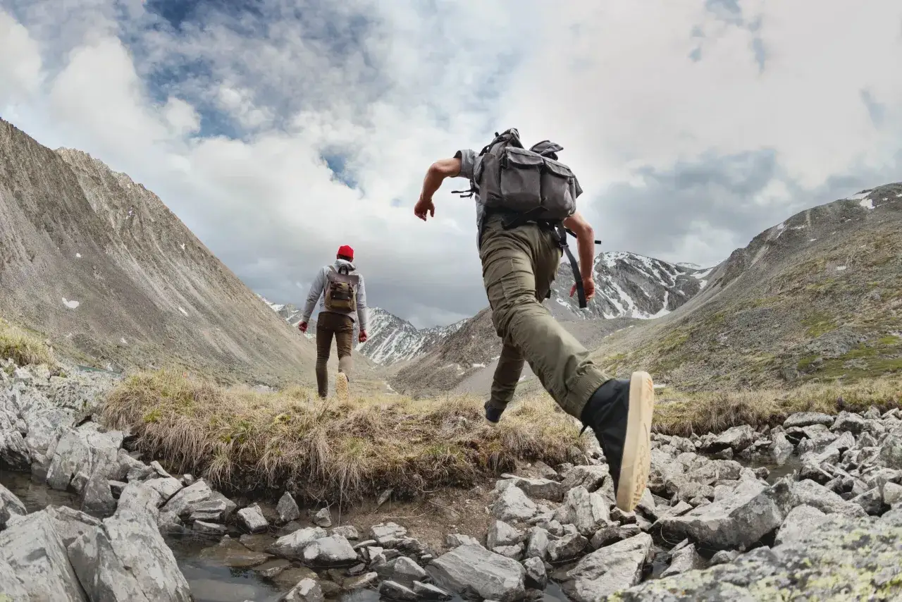 Dwóch turystów wyrusza na bieganie po górach. Jeden skacze przez strumyk, drugi idzie przed nim.