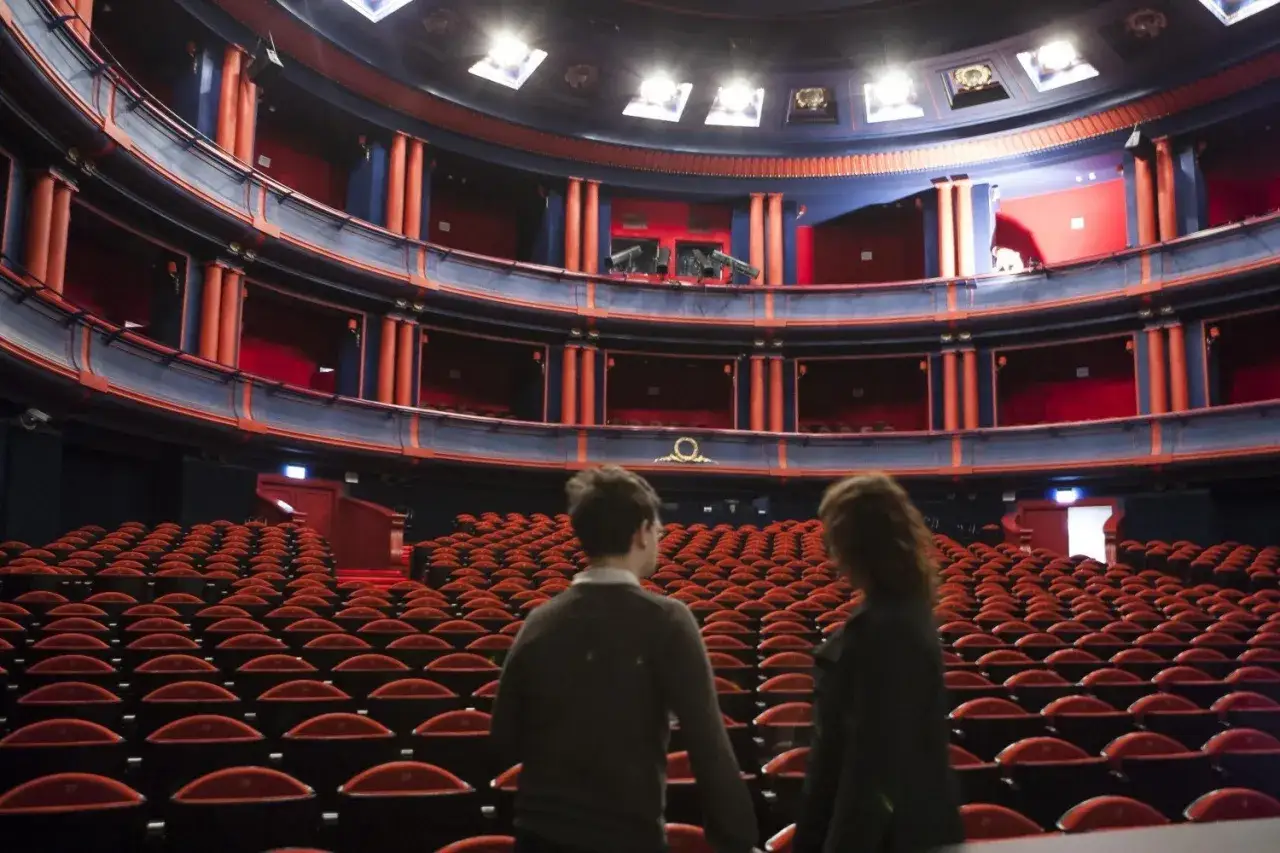 Wnętrze Teatru Roma w Warszawie. Puste czerwone fotele czekają na widzów, a na widowni stoją dwie osoby.