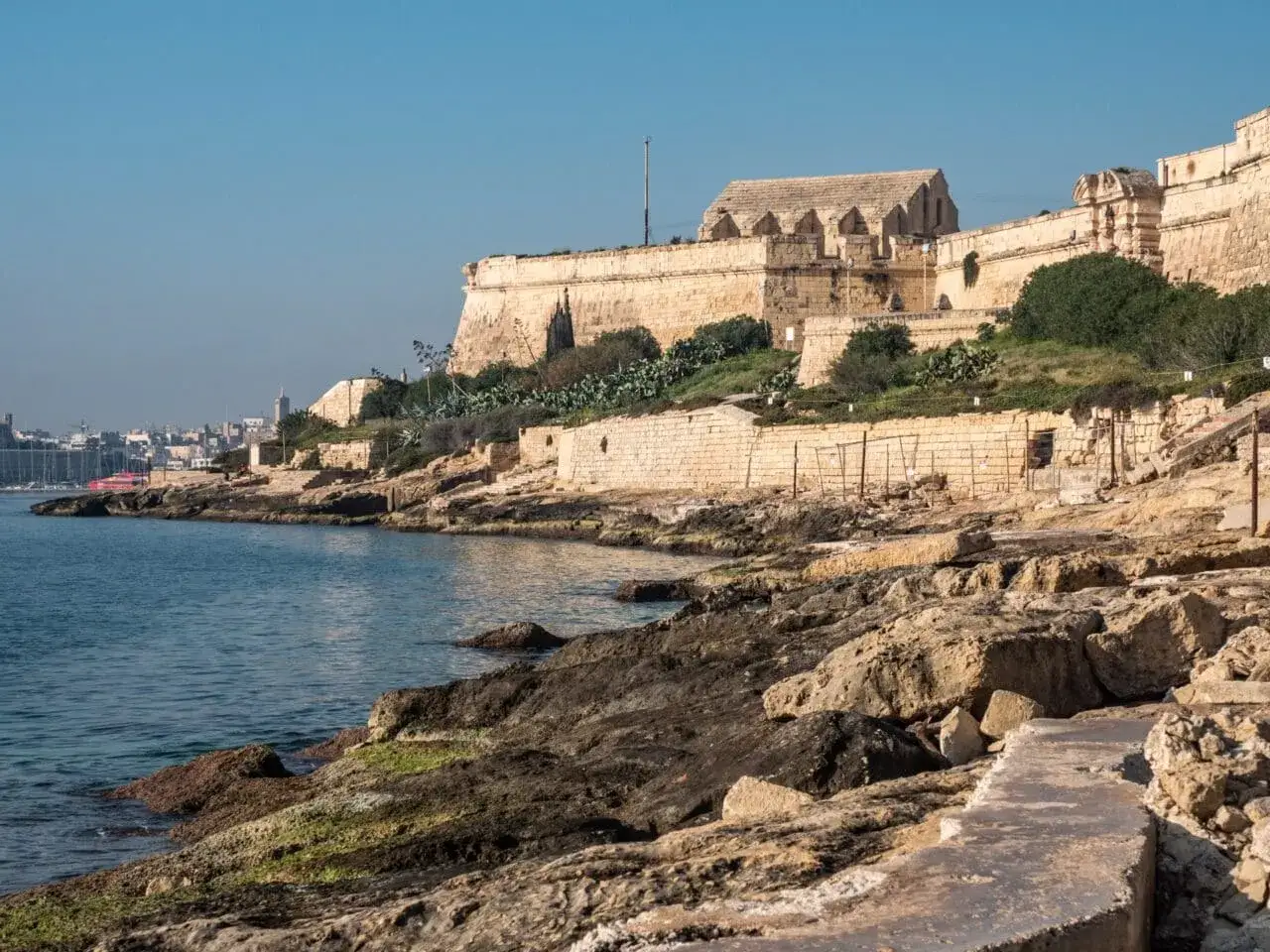 Kamienne fortyfikacje nad lazurowym morzem, z widokiem na miasto w oddali. To właśnie gdzie leży Malta na mapie – śródziemnomorski klejnot.